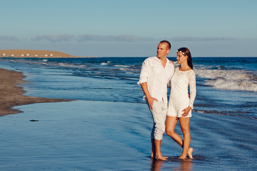 A couple enjoys a romantic walk on a sunny beach with the ocean in the background.