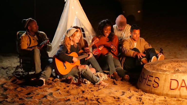 A diverse group of friends playing music around a beach bonfire at night, socializing and having fun.